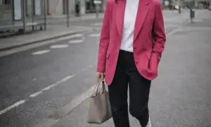Kobieta w różowej marynarce w stylu barbiecore, białej koszulce i jeansach idzie ulicą z szarą torbą.