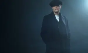 Mężczyzna w kaszkiecie i płaszczu, styl Peaky Blinders, z rękami w kieszeniach, w zadymionym, mrocznym otoczeniu.