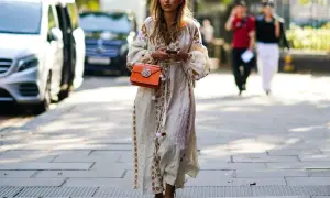 Kobieca stylizacja: jakie buty do sukienki boho? Modelka w zwiewnej, haftowanej sukience i sandałkach na obcasie, z pomarańczową torebką.