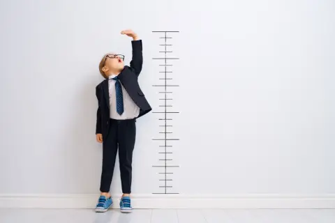 Chłopiec w garniturze mierzy wzrost przy tablicy. Czy ma już rozmiar 92? Jaki wiek?