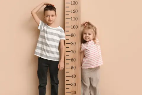 Chłopiec i dziewczynka mierzą wzrost przy drewnianej miarce. Chłopiec ma ok. 115 cm, dziewczynka ok. 85 cm. Rozmiar 68 jaki wiek?