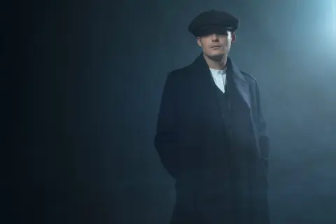 Mężczyzna w kaszkiecie i płaszczu, styl Peaky Blinders, z rękami w kieszeniach, w zadymionym, mrocznym otoczeniu.