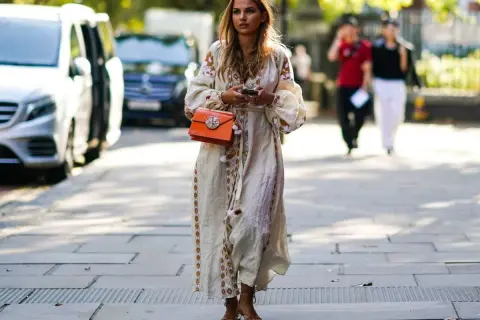 Kobieca stylizacja: jakie buty do sukienki boho? Modelka w zwiewnej, haftowanej sukience i sandałkach na obcasie, z pomarańczową torebką.