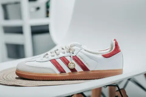 Białe buty Adidas Samba z czerwonymi paskami i zamszowym noskiem, ozdobione złotym medalionem.
