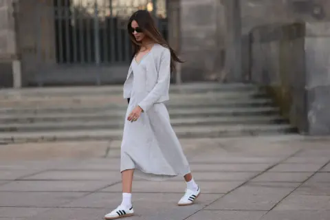 Elegancka sukienka i sportowe buty tworzą stylowy kontrast. Kobieta w szarym komplecie i białych sneakersach idzie ulicą.