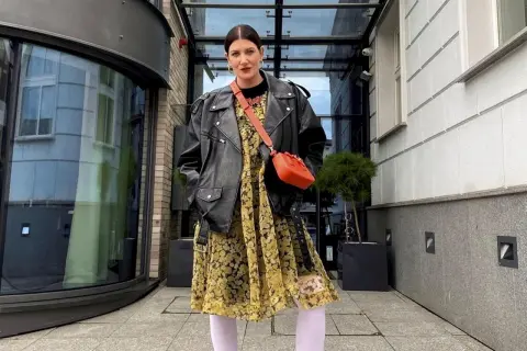 Kobieta w skórzanej ramonesce i żółtej sukience w kwiaty, z pomarańczową torebką na ramieniu.