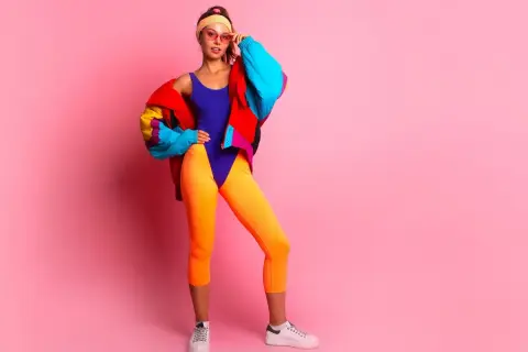 Dziewczyna w neonowym stroju i kolorowej kurtce, z opaską na włosach i okularach, prezentuje styl lat 90.