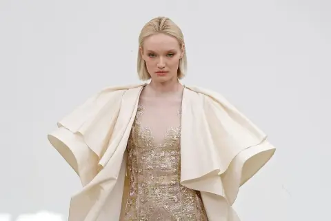 Modelka w kremowym, falbaniastym płaszczu i zdobionej sukni prezentuje dzieło haute couture.