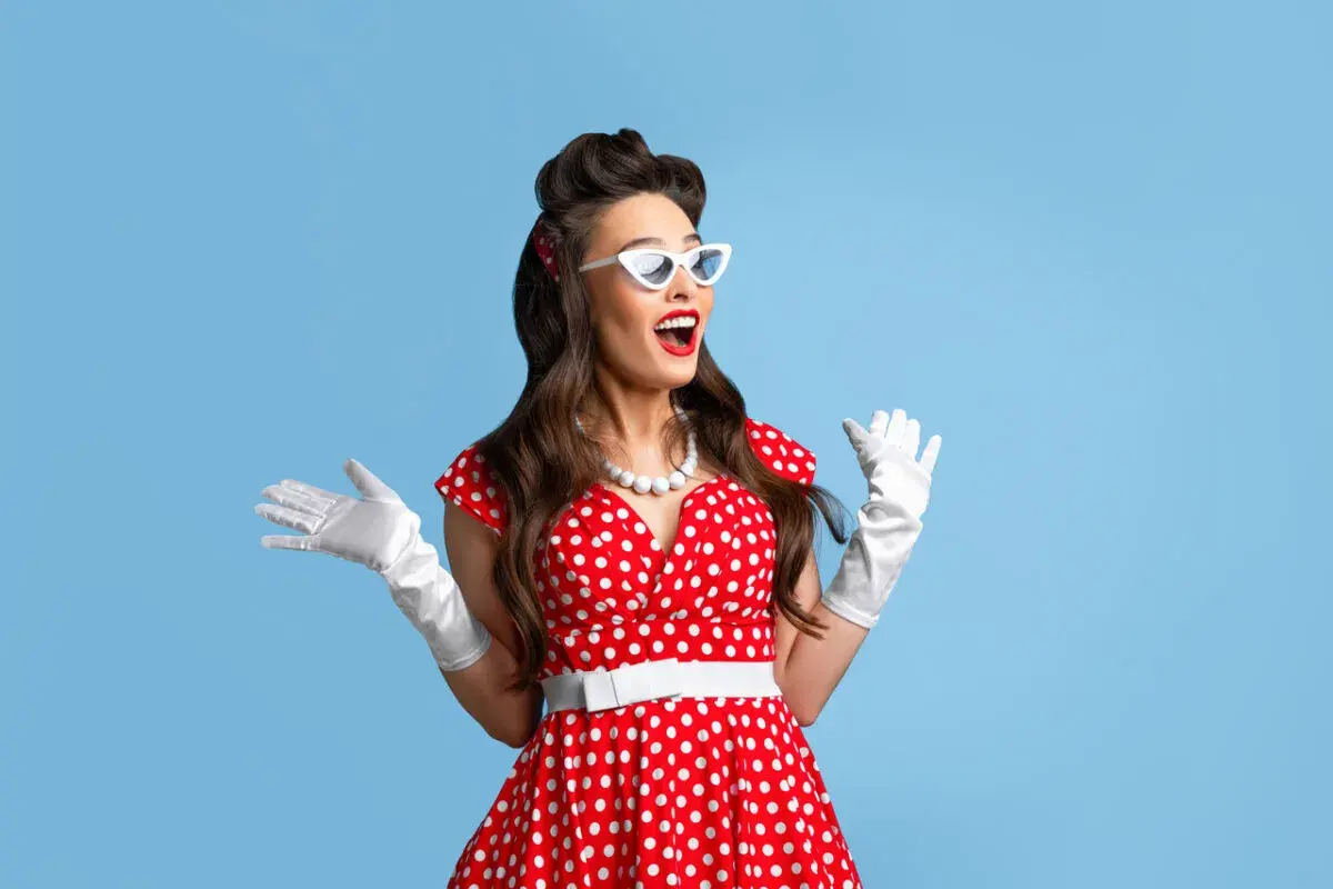 Kobieta w czerwonej sukience w grochy, białych rękawiczkach i okularach w stylu pin up girl stylizacje, z uniesionymi rękami.