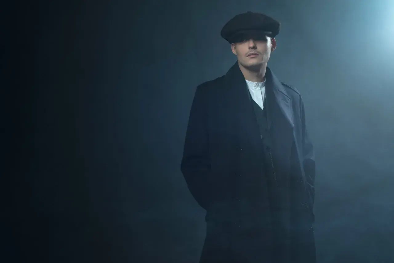Mężczyzna w kaszkiecie i płaszczu, styl Peaky Blinders, z rękami w kieszeniach, w zadymionym, mrocznym otoczeniu.