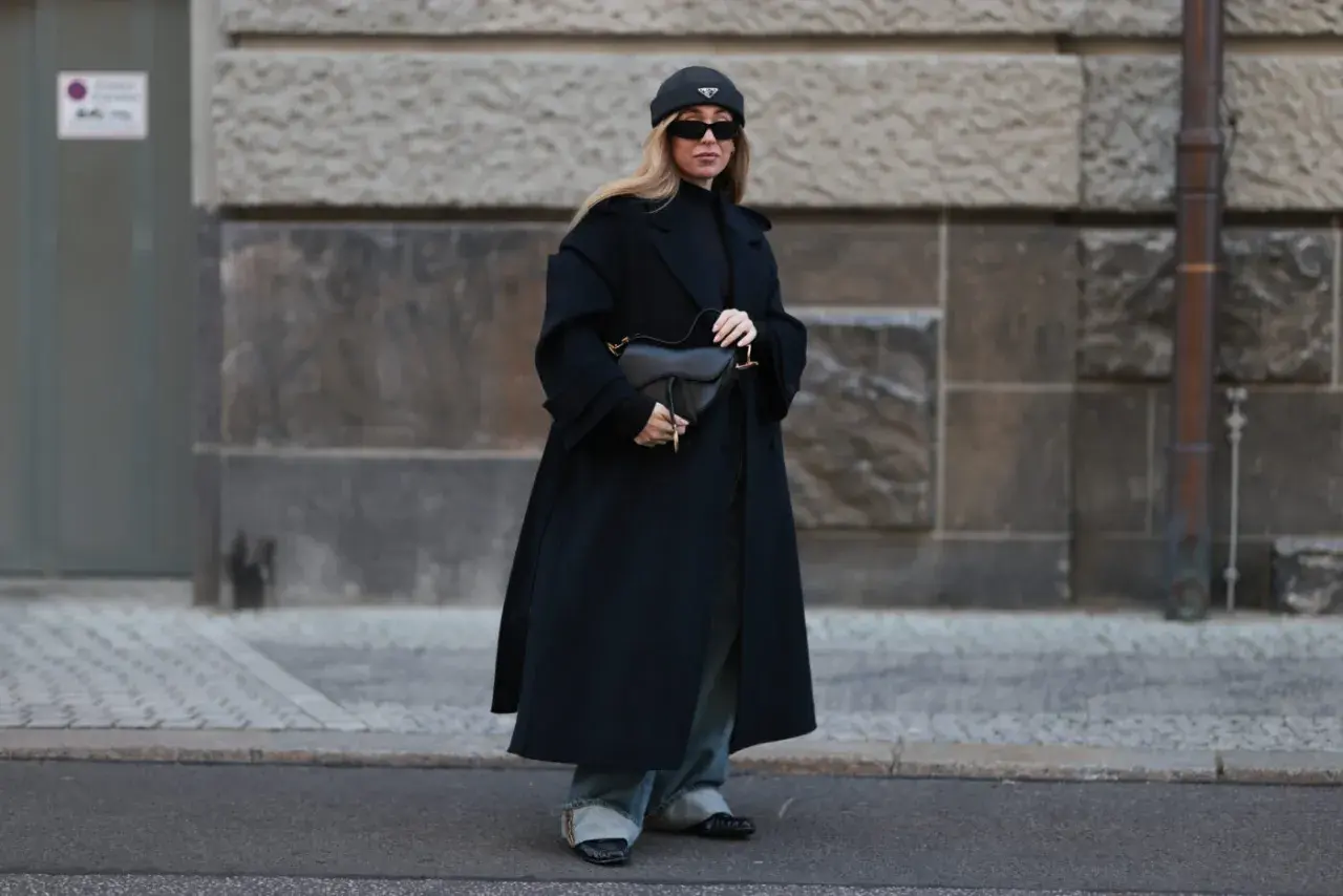 Elegancka stylizacja płaszcz i adidasy. Kobieta w czarnym płaszczu, jeansach i czapce z daszkiem, trzyma torebkę.
