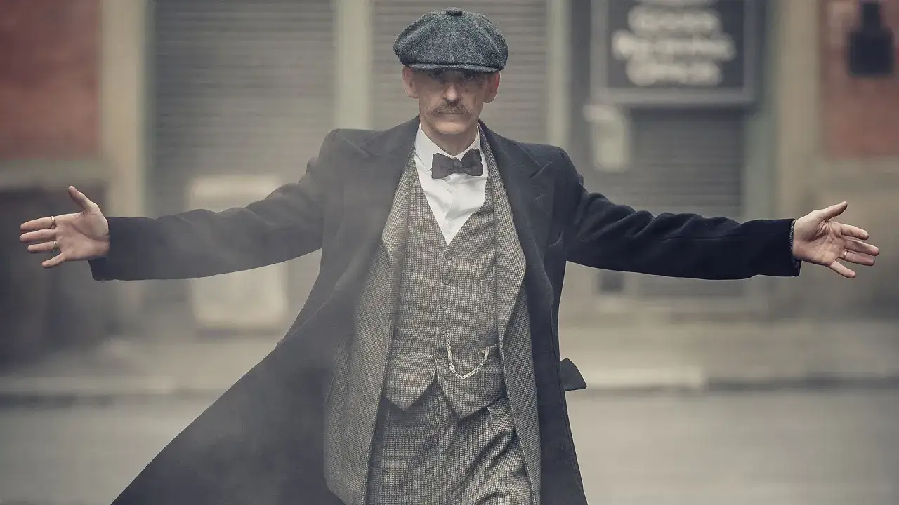 Mężczyzna w czapce i płaszczu, w stylu Peaky Blinders, z rozłożonymi ramionami.