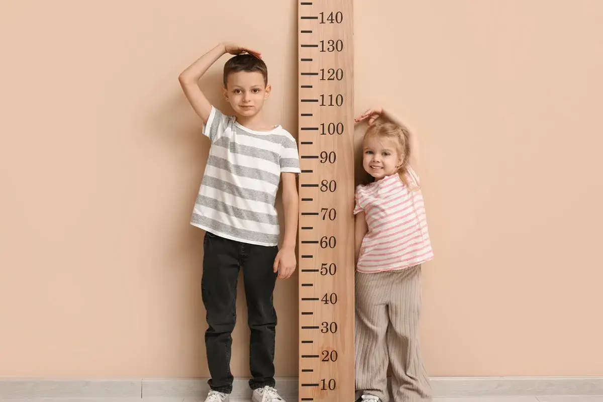 Chłopiec i dziewczynka mierzą wzrost przy drewnianej miarce. Chłopiec ma ok. 134 cm, co sugeruje wiek 8-9 lat.