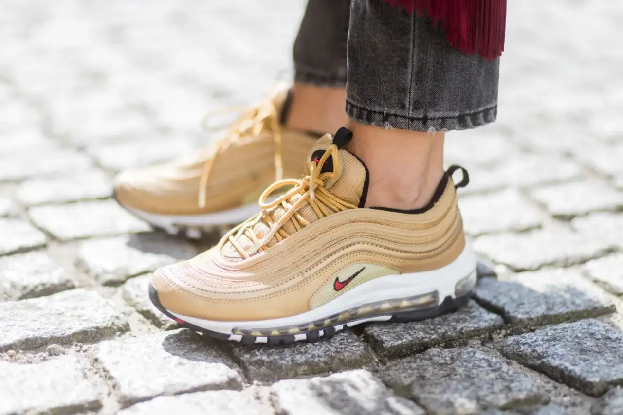 Złote sneakersy Nike Air Max 97, idealne do miejskich stylizacji młodzieżowych, na kamiennej kostce brukowej.