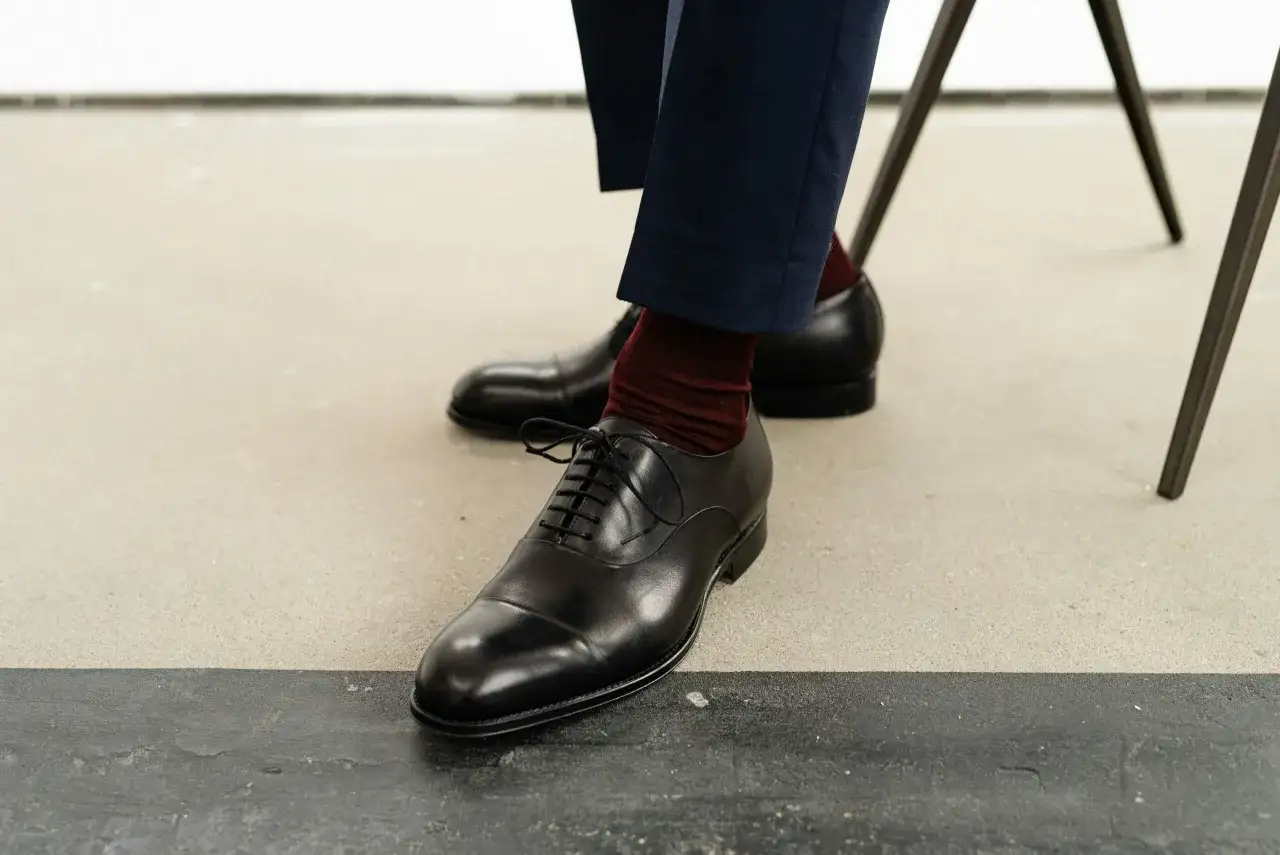 Eleganckie, czarne, sznurowane buty męskie z bordowymi skarpetkami. Klasyczny, sartorialny styl podkreślony przez granatowe spodnie.