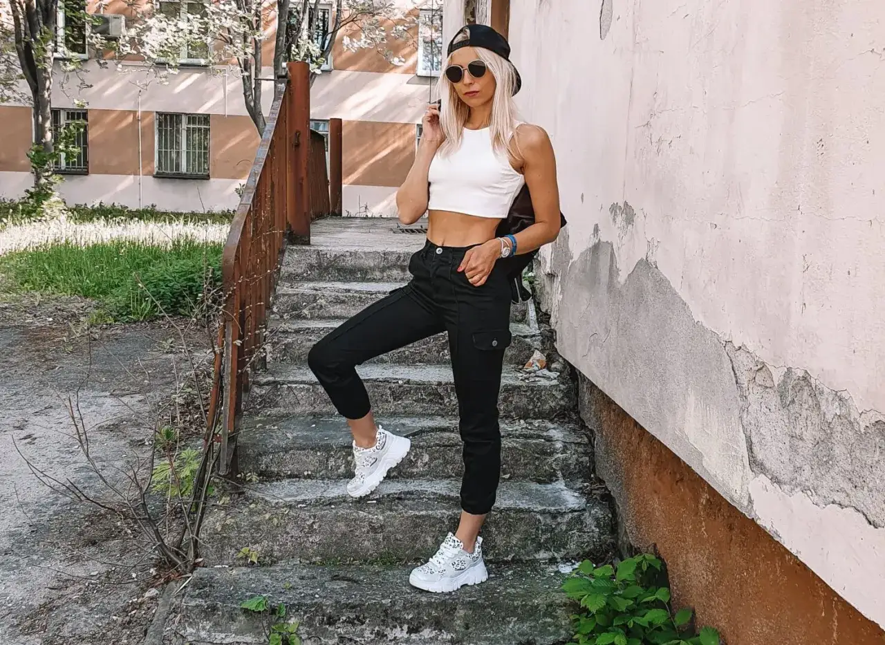 Kobieta w białym topie i czarnych spodniach cargo, na nogach ma białe buty na grubej podeszwie, zwane sneakersami typu chunky.