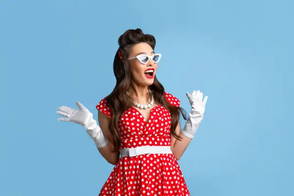 Kobieta w czerwonej sukience w grochy, białych rękawiczkach i okularach w stylu pin up girl stylizacje, z uniesionymi rękami.