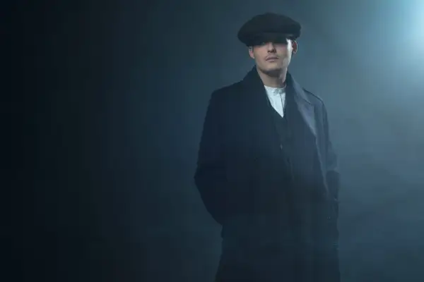 Mężczyzna w kaszkiecie i płaszczu, styl Peaky Blinders, z rękami w kieszeniach, w zadymionym, mrocznym otoczeniu.