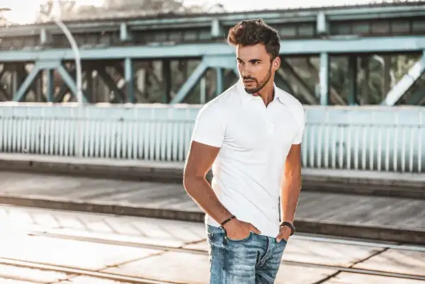 Mężczyzna w białej koszulce polo i jeansach stoi na tle mostu.