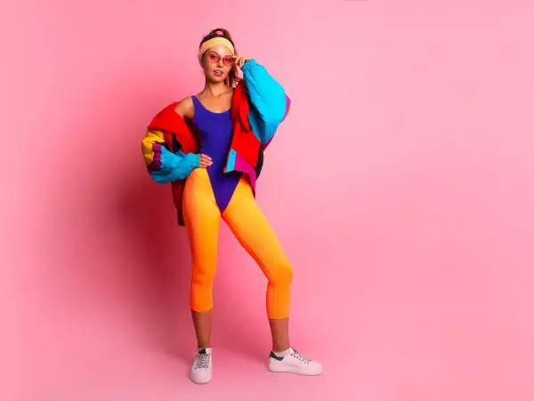 Dziewczyna w neonowym stroju i kolorowej kurtce, z opaską na włosach i okularach, prezentuje styl lat 90.