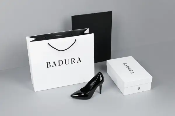 Elegancki czarny szpilka od firmy Badura, obok białej torby i pudełka z logo. Badura to polska marka obuwia.
