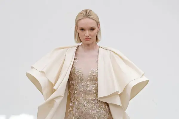 Modelka w kremowym, falbaniastym płaszczu i zdobionej sukni prezentuje dzieło haute couture.