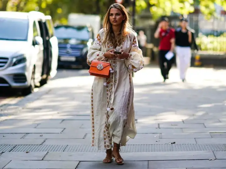 Kobieca stylizacja: jakie buty do sukienki boho? Modelka w zwiewnej, haftowanej sukience i sandałkach na obcasie, z pomarańczową torebką.